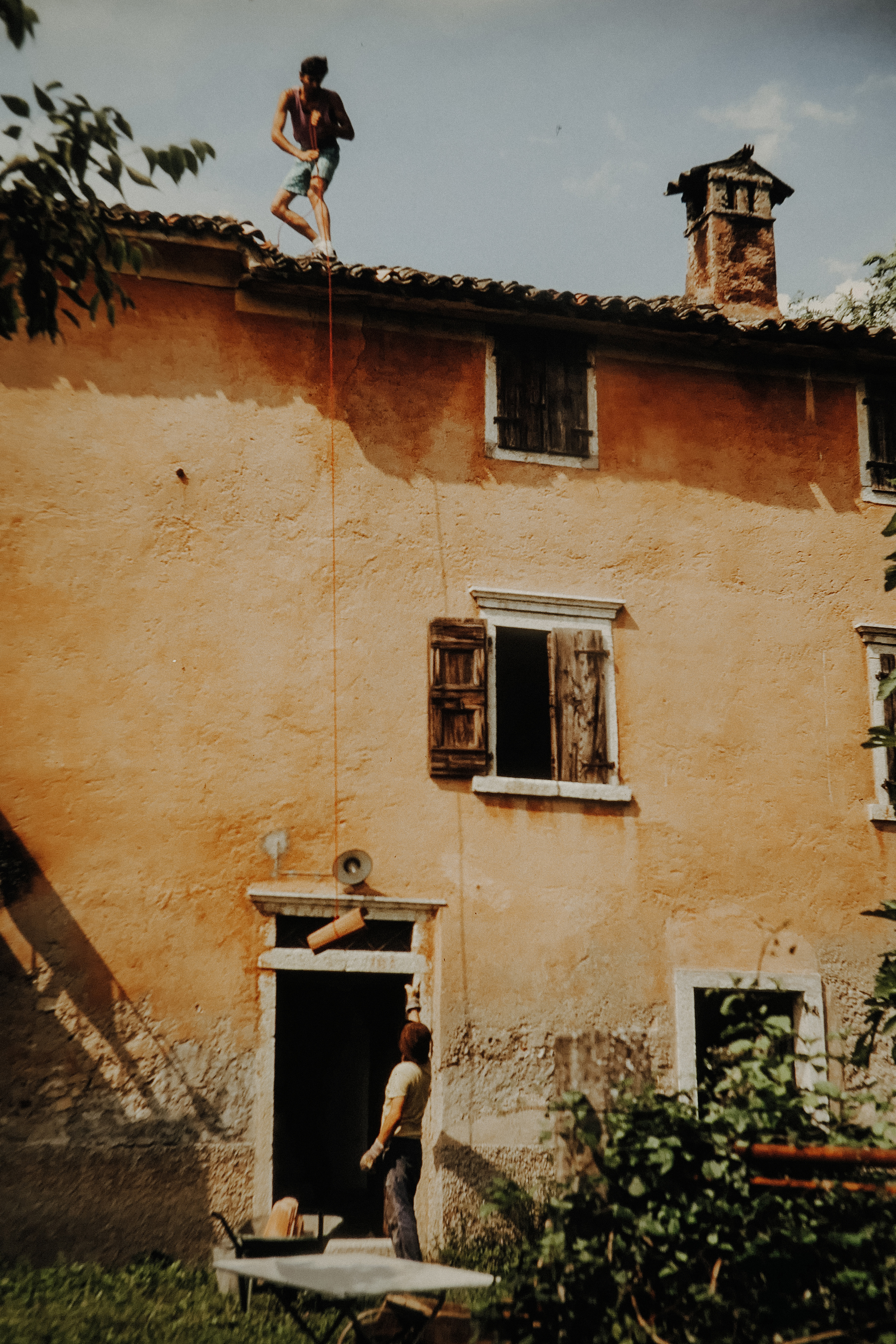 Eine Person steht auf dem Dach und lässt per Seil Ziegelsteine herunter – eine Person nimmt es am Boden entgegen.